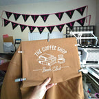 Brown bag with 'The Coffee Shop Book Club' design held by a hand, with a banner and printer in the background.