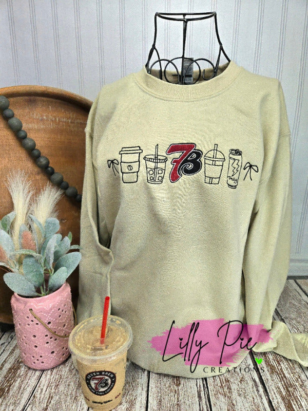 Beige sweatshirt with coffee cup graphics on a wooden surface with a plant and cup.