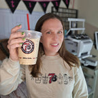 Person holding a coffee cup with a branded design, wearing a matching sweatshirt in a casual setting.