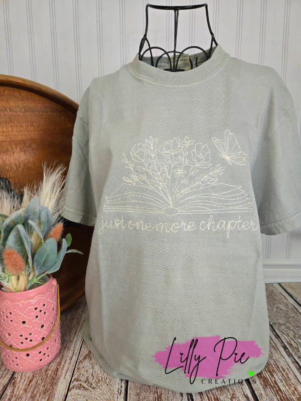 sage t-shirt with floral design and text 'just one more chapter' on a wooden surface with decorative items.