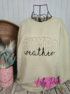 Beige sweatshirt with 'Gumbo weather' text on a wooden surface with decorative elements.