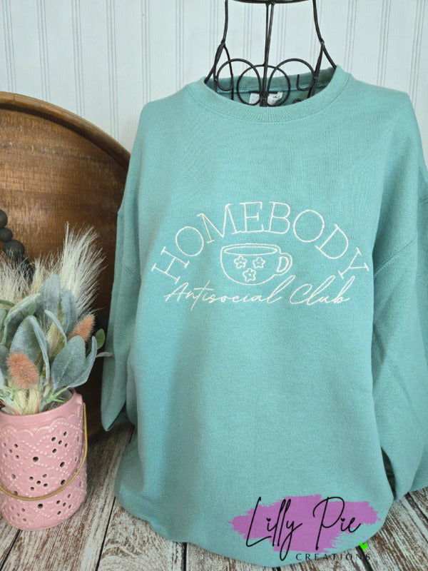 Teal sweatshirt with 'Homebody Antisocial Club' text on a wooden surface with decorative items.