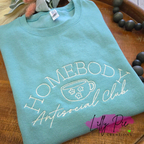 Light blue sweatshirt with 'Homebody Antisocial Club' text and a cup graphic on a wooden surface.