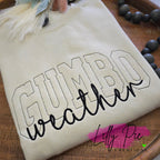 Beige sweatshirt with 'Gumbo weather' text on a wooden surface, featuring the brand 'Lilly Pie Creations'.