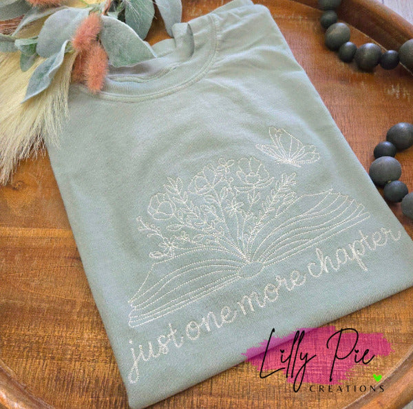 Sage shirt with floral book design Just one more chapter text on a wooden surface, featuring the brand 'Lilly Pre Creations'.