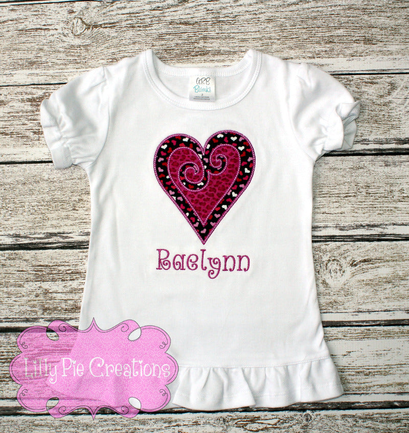A white short-sleeved girls' shirt with a heart applique in pink and black colors, and the name 'Baylinn' embroidered on it.